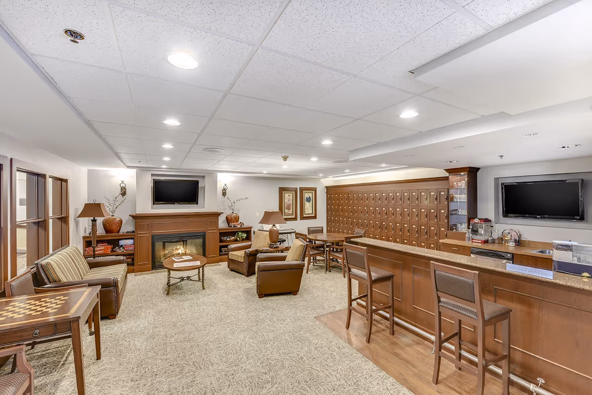 A cozy common area in a senior living facility featuring a fireplace with a TV mounted above it, comfortable seating including armchairs and a striped sofa, a wooden table with a chessboard, a counter with bar stools, and a wall of mailboxes. The room is well-lit with recessed ceiling lights and decorated with lamps, vases, and framed artwork.