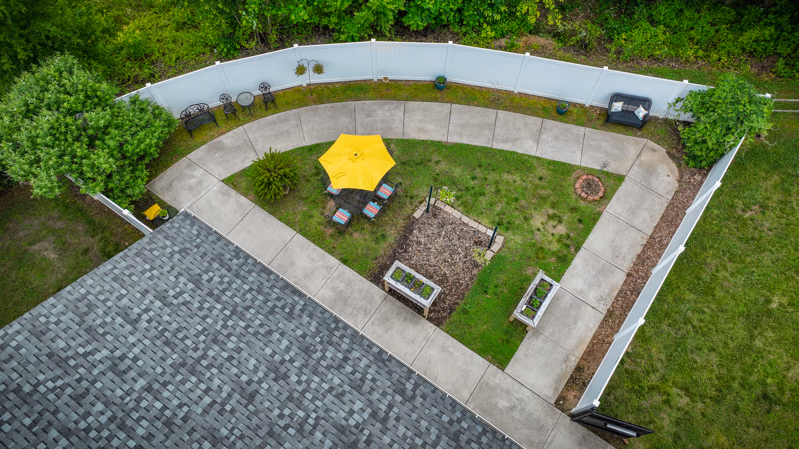 Aerial view of a fenced courtyard with a concrete walkway, seating around a yellow umbrella, planters and surrounding greenery.