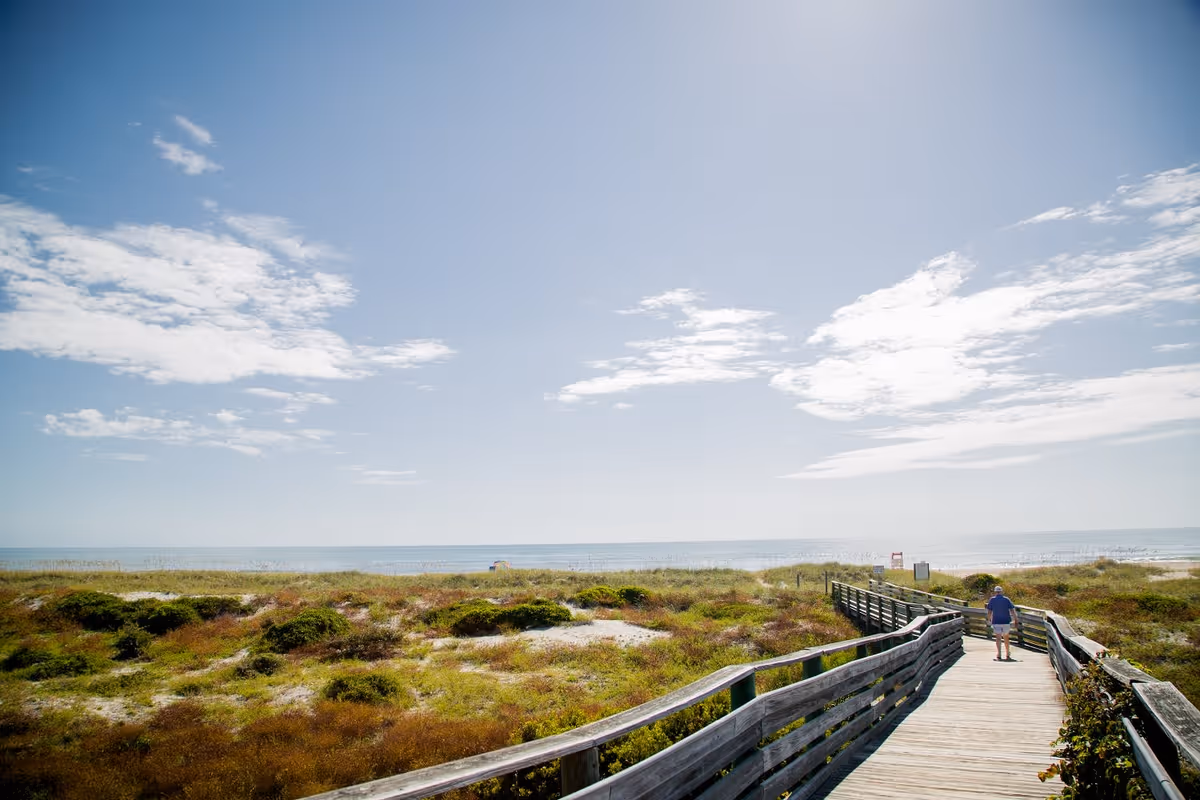 A wooden boardwalk leading through grassy dunes towards a beach with the ocean visible in the background under a partly cloudy blue sky. A person is walking on the boardwalk towards the beach.