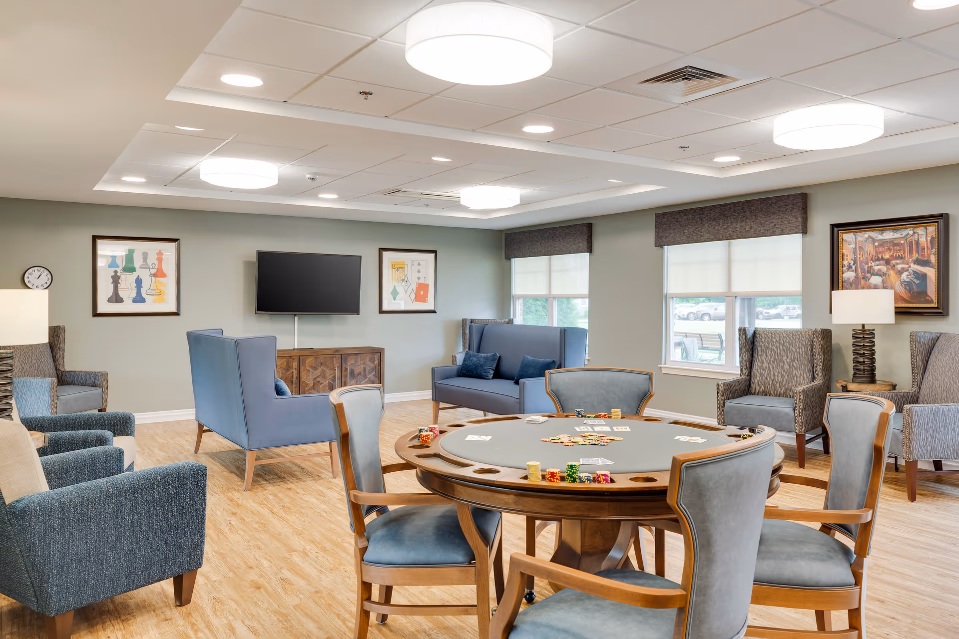 A well-lit common room with a round poker table in the center, surrounded by six chairs. The table has poker chips and playing cards on it. The room features multiple comfortable armchairs and sofas arranged around the perimeter. There is a flat-screen TV mounted on the wall, framed artwork, and two windows with blinds partially drawn. The floor is wooden, and the ceiling has recessed lighting and round ceiling lights.