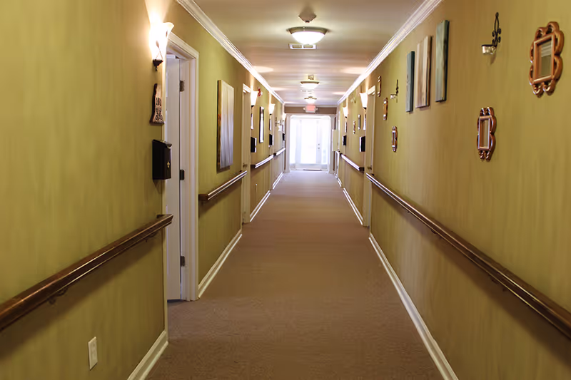 Long carpeted corridor in a care home with handrails, wall sconces, and wall decorations leading to an exit door.