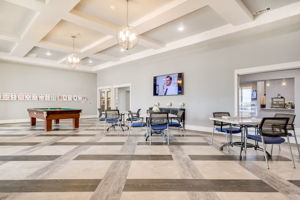 Large senior living common room with a pool table, multiple tables and chairs, and a wall-mounted TV.
