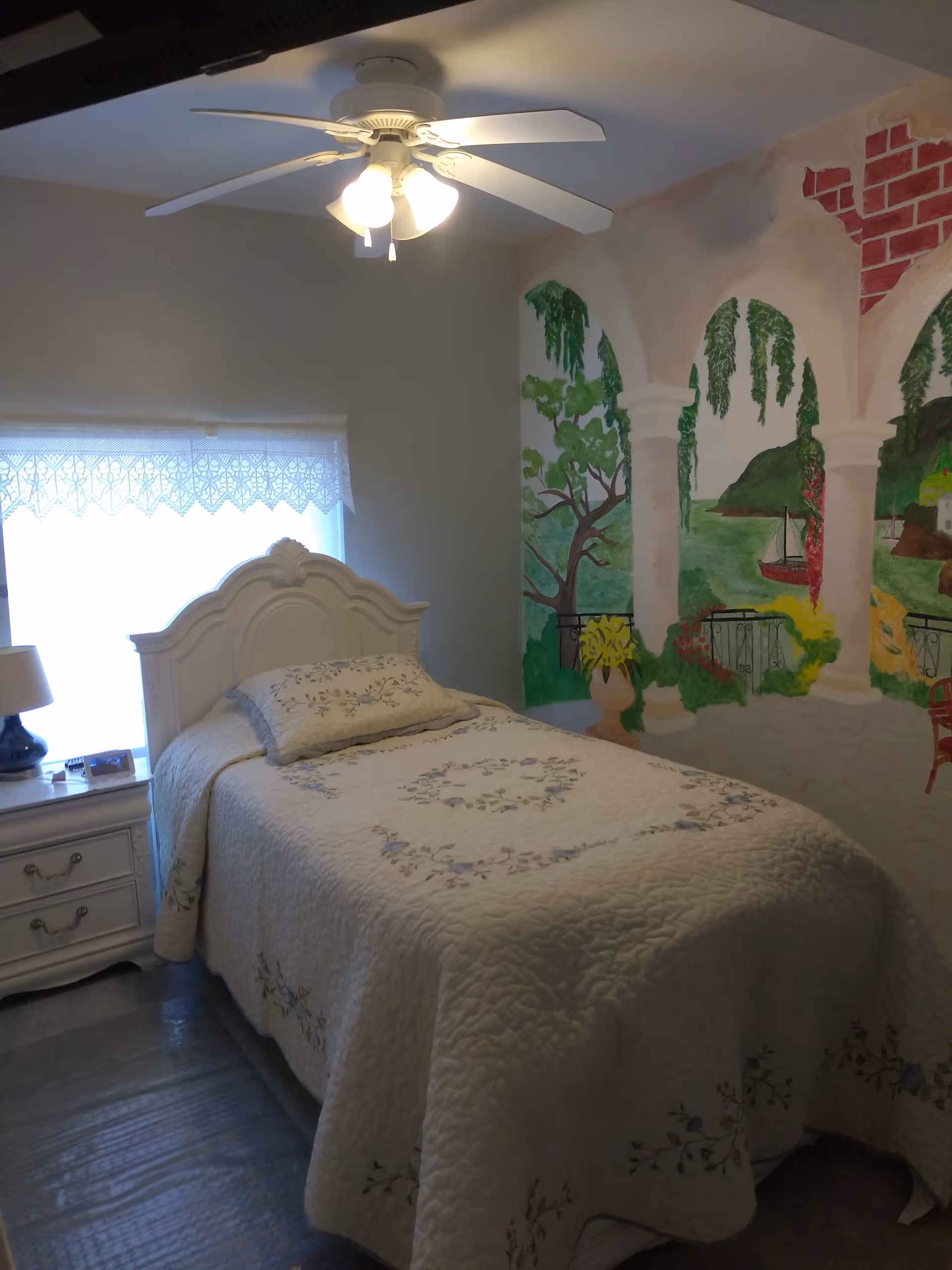A cozy single bedroom with a white bed, bedside table, ceiling fan, lace-curtained window and a colorful mural on one wall.