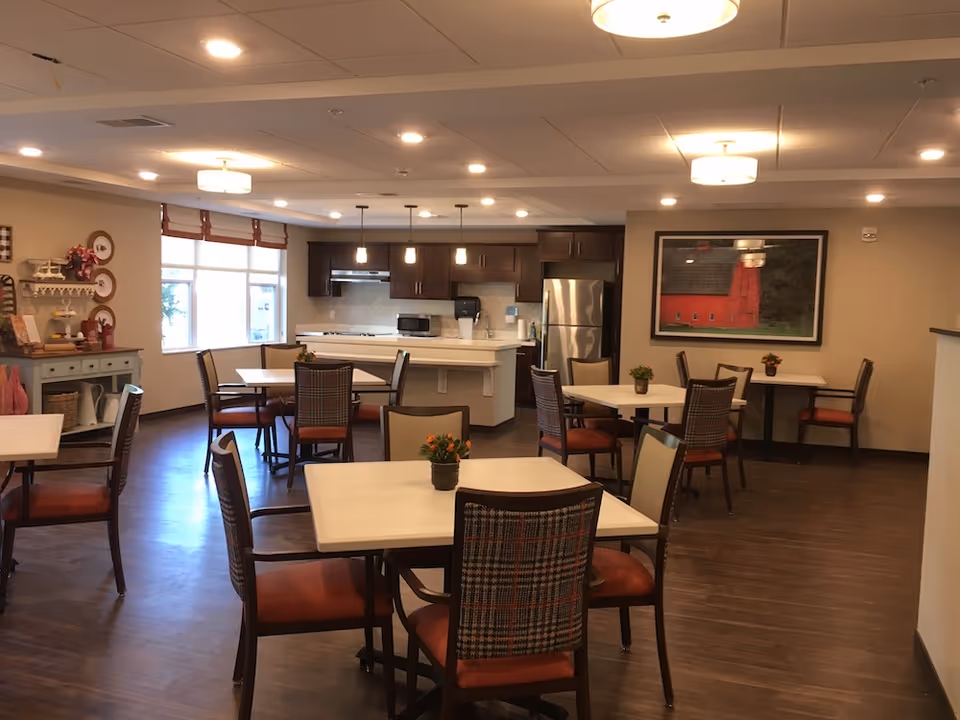 Communal dining area with multiple tables and chairs and a kitchen in the background.