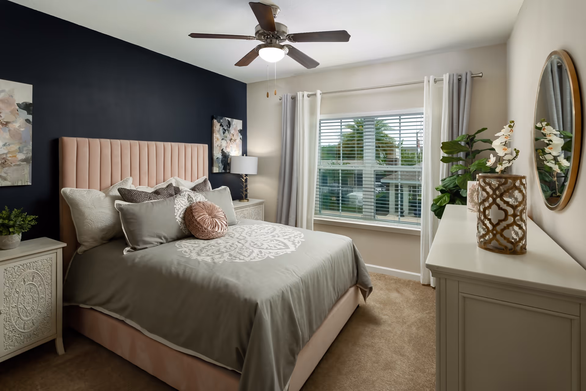 A cozy bedroom with a large bed featuring a pink upholstered headboard and gray bedding with decorative pillows. The room has a dark accent wall behind the bed, two floral paintings, a nightstand with a lamp, a window with white curtains and blinds, and a white dresser with a round mirror and decorative items including a plant and a vase.