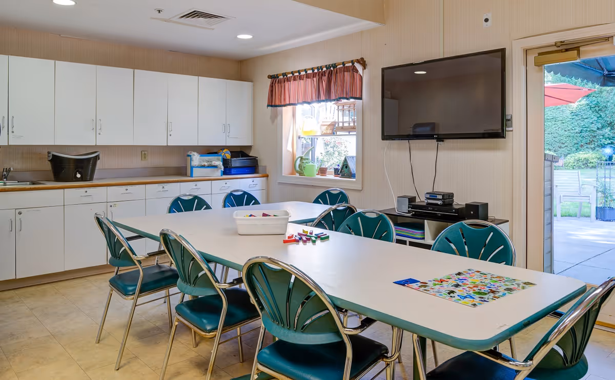 Activity/dining room with a long table surrounded by teal chairs, cabinets along the wall, a wall-mounted TV, and a door leading outside.