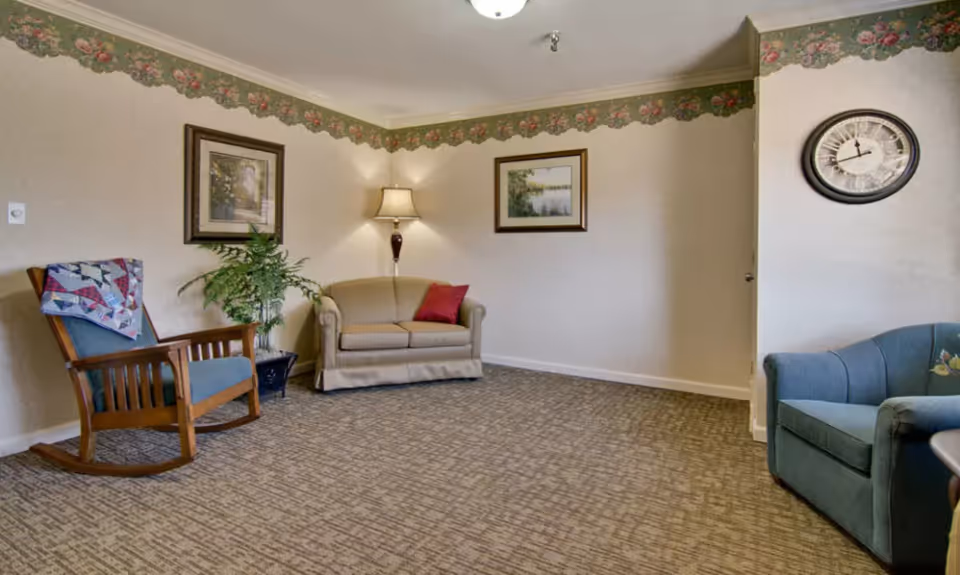 A cozy living room with a beige loveseat, a wooden rocking chair with a quilt draped over it, a blue armchair, a floor lamp, two framed pictures on the walls, a round wall clock, and a potted plant. The walls have a floral wallpaper border near the ceiling.