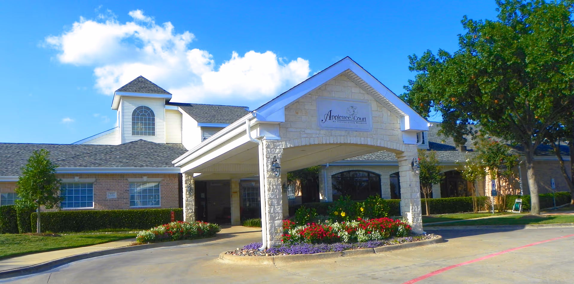 Front entrance of Appletree Court with a covered porte-cochère, landscaped flowerbeds, and a brick-and-stone facade under a blue sky.