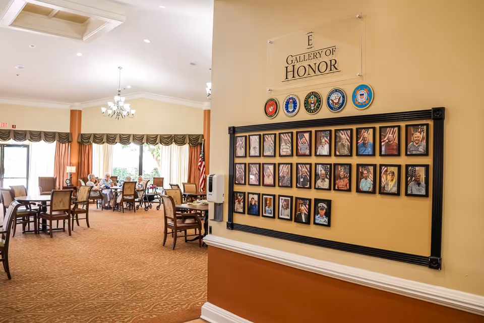 Interior view of a senior living facility dining area with several elderly residents seated at tables. On the right wall, there is a 'Gallery of Honor' display featuring framed photos of veterans and military service emblems above it. The room is well-lit with large windows and chandeliers.