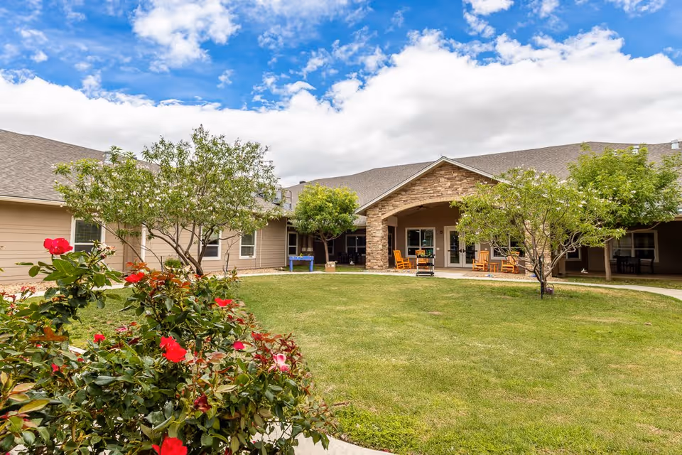 A courtyard area at Bristol Park at Amarillo Assisted & Living Memory Care featuring a well-maintained lawn, flowering bushes with red flowers, several small trees, and a covered patio with rocking chairs. The building surrounds the courtyard with beige siding and a stone accent on the patio entrance. The sky is partly cloudy with patches of blue.