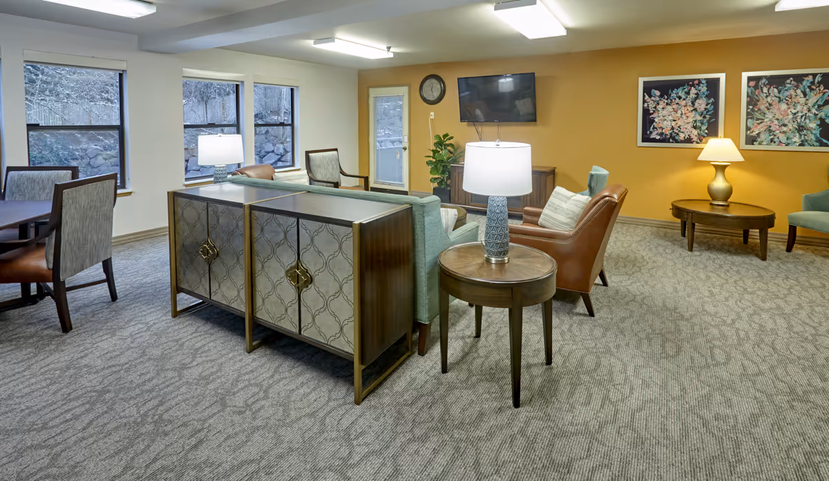 A cozy senior living common area with a mix of seating including armchairs and a sofa. There are two side tables with lamps, a wall-mounted flat screen TV, a clock, and two framed floral artworks on a mustard yellow accent wall. The room has carpeted flooring and several windows letting in natural light.