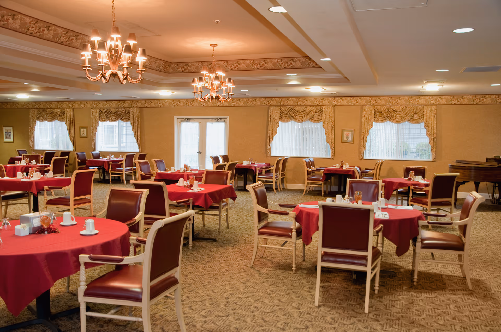 Spacious dining room with round tables draped in red tablecloths, upholstered chairs, chandeliers, and curtained windows.