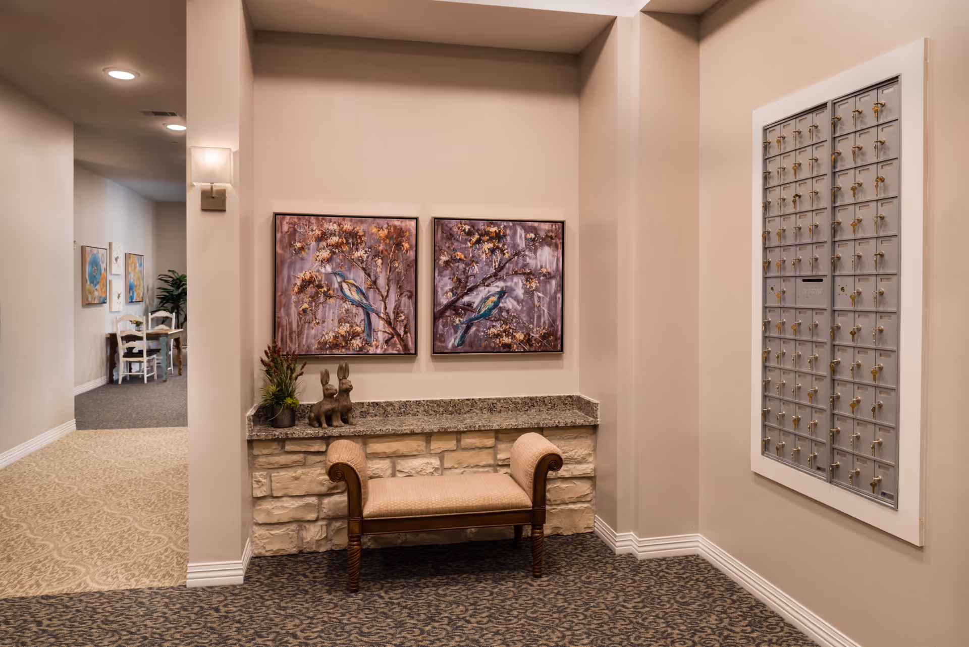 Interior hallway of a senior living facility with a cushioned bench against a stone and granite ledge. Above the bench are two framed paintings of birds on branches. To the right, there is a wall-mounted mailbox unit with multiple locked compartments. In the background, a small table with chairs and additional wall art is visible.