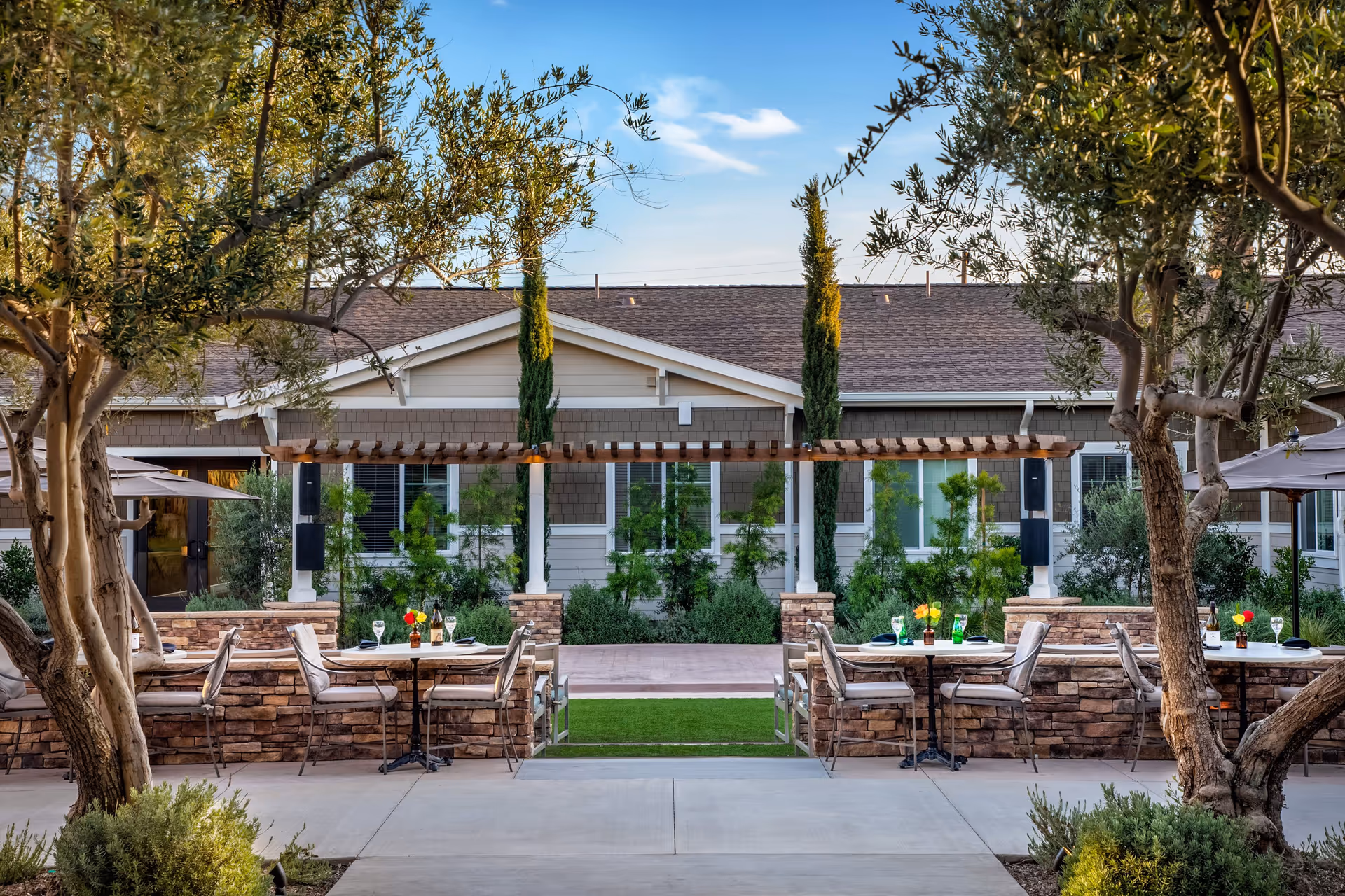 Outdoor patio area with tables and chairs set for dining, surrounded by trees and greenery, with a building in the background under a clear blue sky.