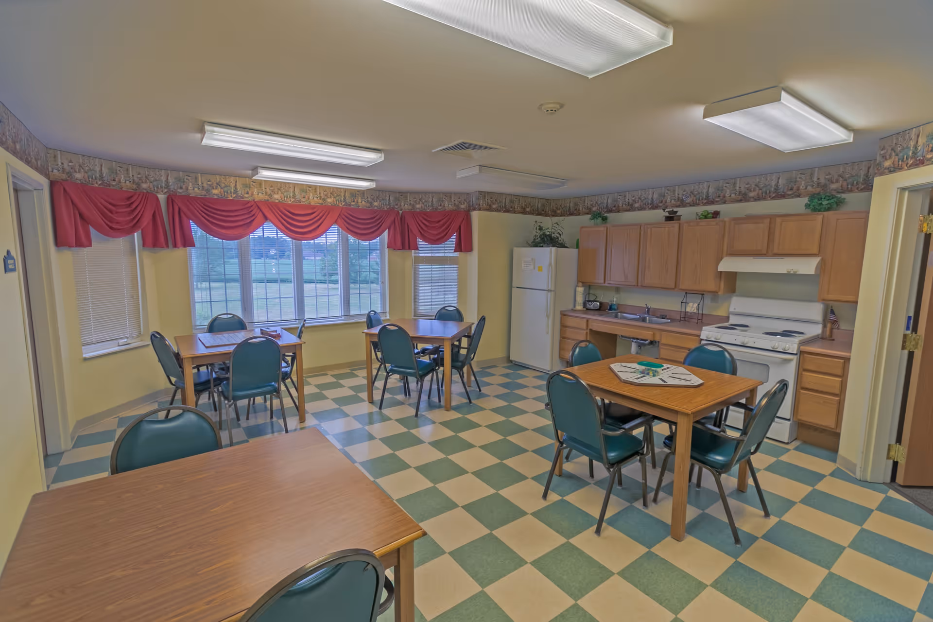 A communal dining and kitchen area with several wooden tables and green chairs arranged on a checkered green and beige floor. The kitchen area includes wooden cabinets, a white refrigerator, a white stove with a range hood, and a sink. Large windows with red valance curtains let in natural light, and fluorescent ceiling lights illuminate the room.