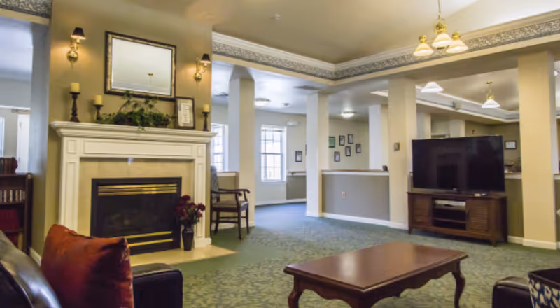 A cozy senior living common area featuring a fireplace with candles and a mirror above it, a large flat-screen TV on a wooden stand, a coffee table, and several chairs. The room has carpeted floors, decorative ceiling trim, and soft lighting.