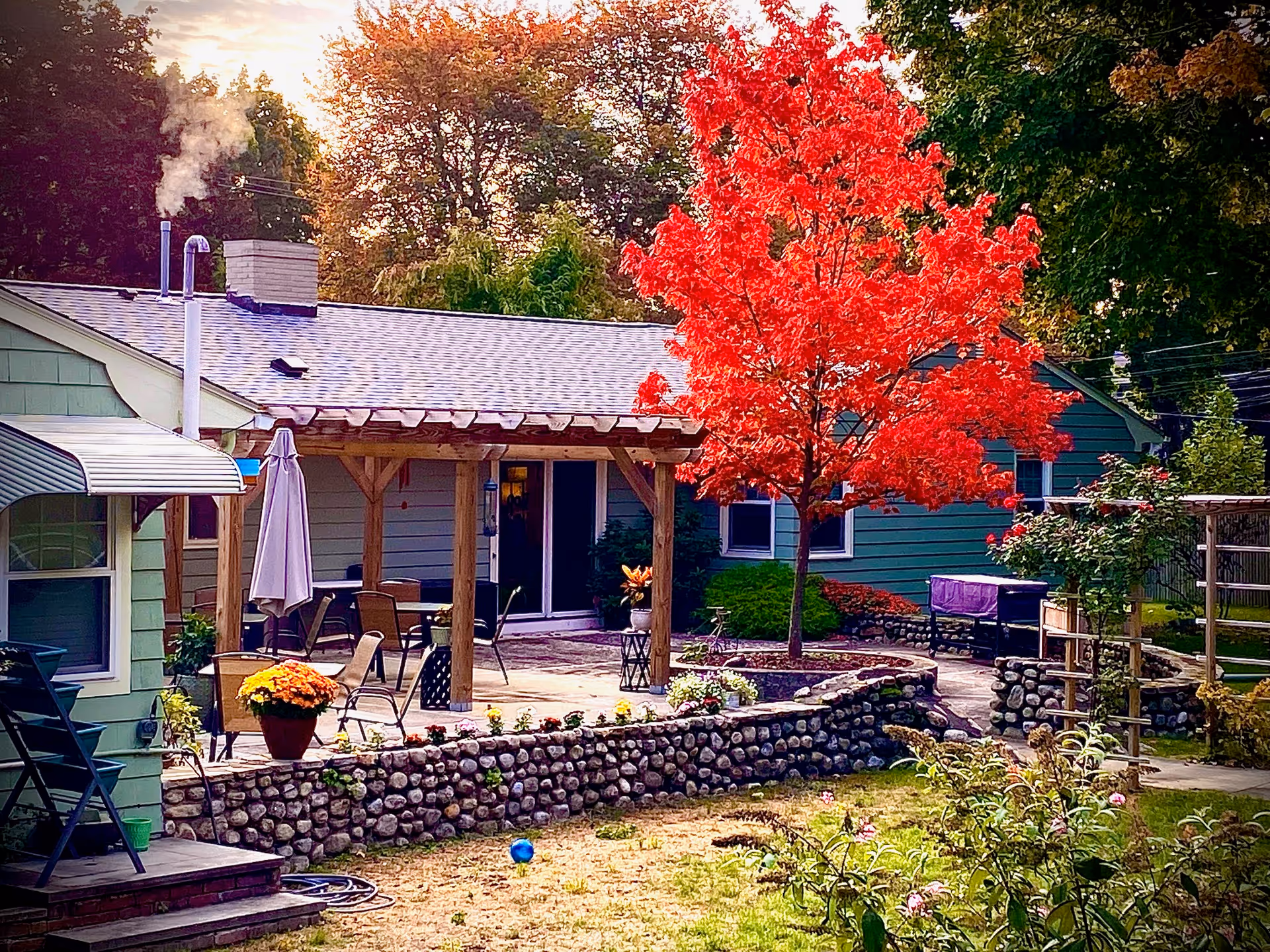 Outdoor patio area of Commonwealth House Assisted Living with a stone wall, several chairs and tables under a wooden pergola, a vibrant red tree, and surrounding greenery in an autumn setting.