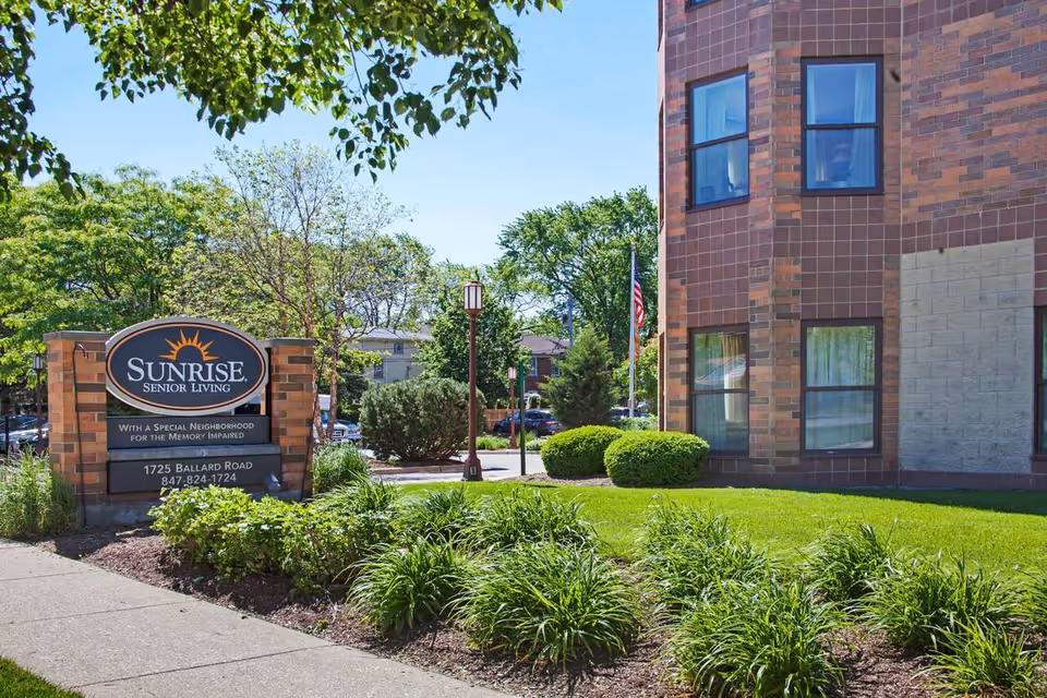 Exterior view of Sunrise Senior Living facility with a brick building, green lawn, bushes, trees, and a sign displaying the facility name and contact information.