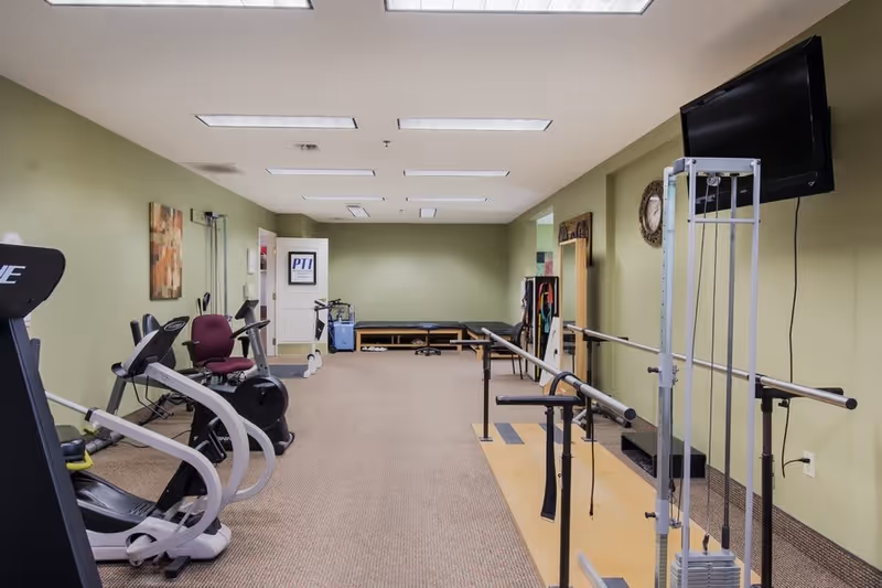 A fitness and rehabilitation room with exercise equipment including stationary bikes, parallel bars for walking support, a weight machine, and a wall-mounted TV. The room has green walls, beige carpet, and fluorescent ceiling lights.