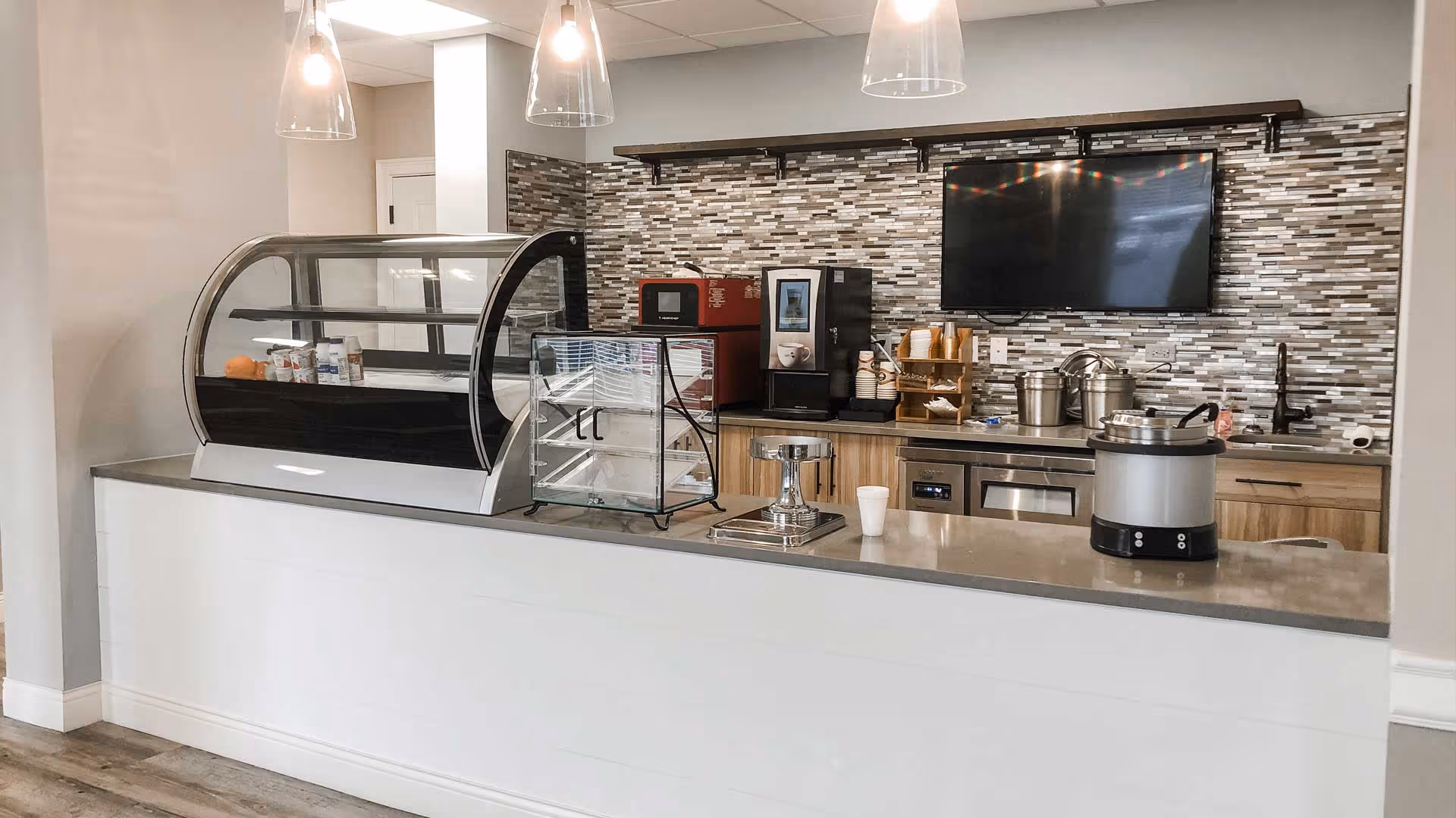 A modern kitchen or serving area with a countertop, glass display cases, a coffee machine, microwave, and a large flat-screen TV mounted on a tiled backsplash wall. The area is well-lit with hanging pendant lights and has wooden cabinets below the counter.
