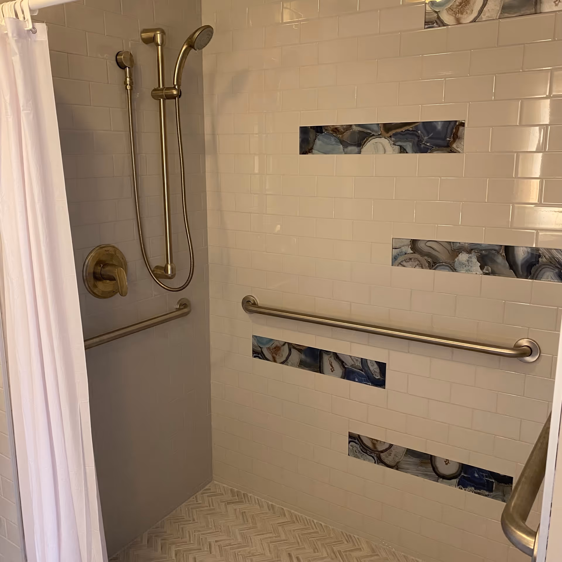 Shower area with white tiled walls featuring decorative blue and gray stone-like tile inserts, a handheld showerhead with a sliding bar, and multiple stainless steel grab bars for support. A white shower curtain is partially visible on the left side.