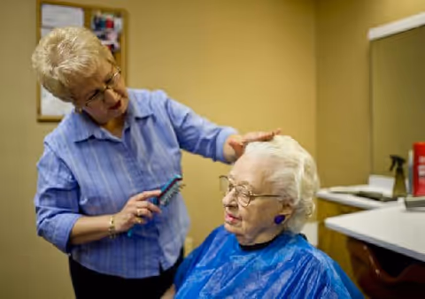An elderly woman with white hair and glasses is seated wearing a blue cape while another woman with short blonde hair and glasses combs her hair in a salon-like setting.