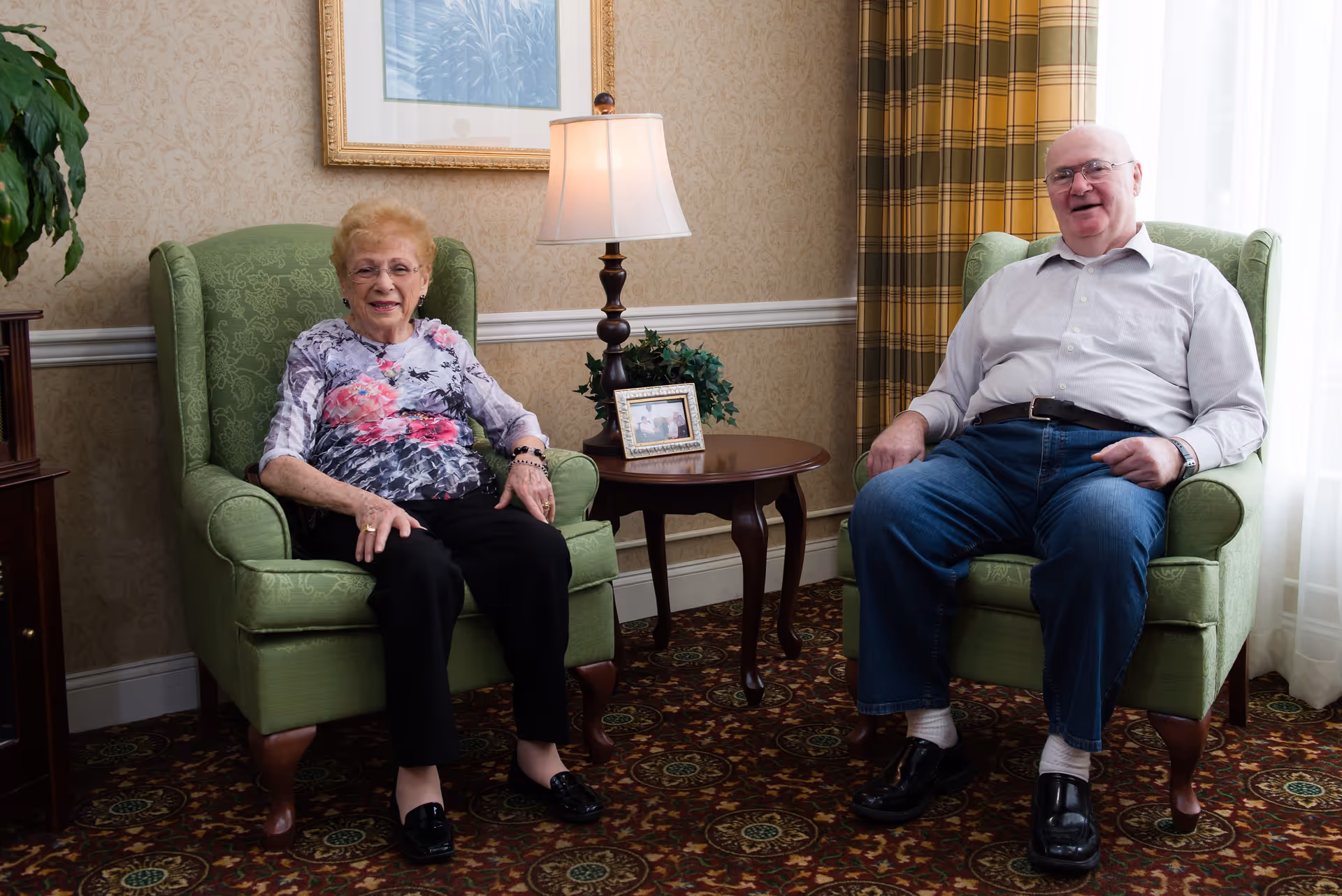 An elderly woman and an elderly man sitting comfortably in green armchairs in a warmly decorated living room with patterned carpet, a side table with a lamp and framed photo, a plant, and a window with plaid curtains.