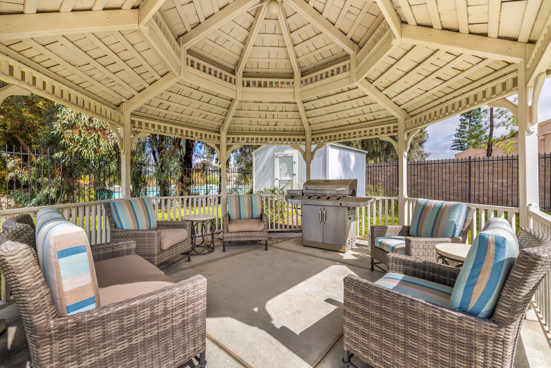 Outdoor gazebo with a white wooden roof and railing, furnished with wicker chairs and sofas featuring striped cushions in blue, beige, and brown tones. There are small round tables and a stainless steel barbecue grill inside the gazebo. Trees and a fence are visible in the background.