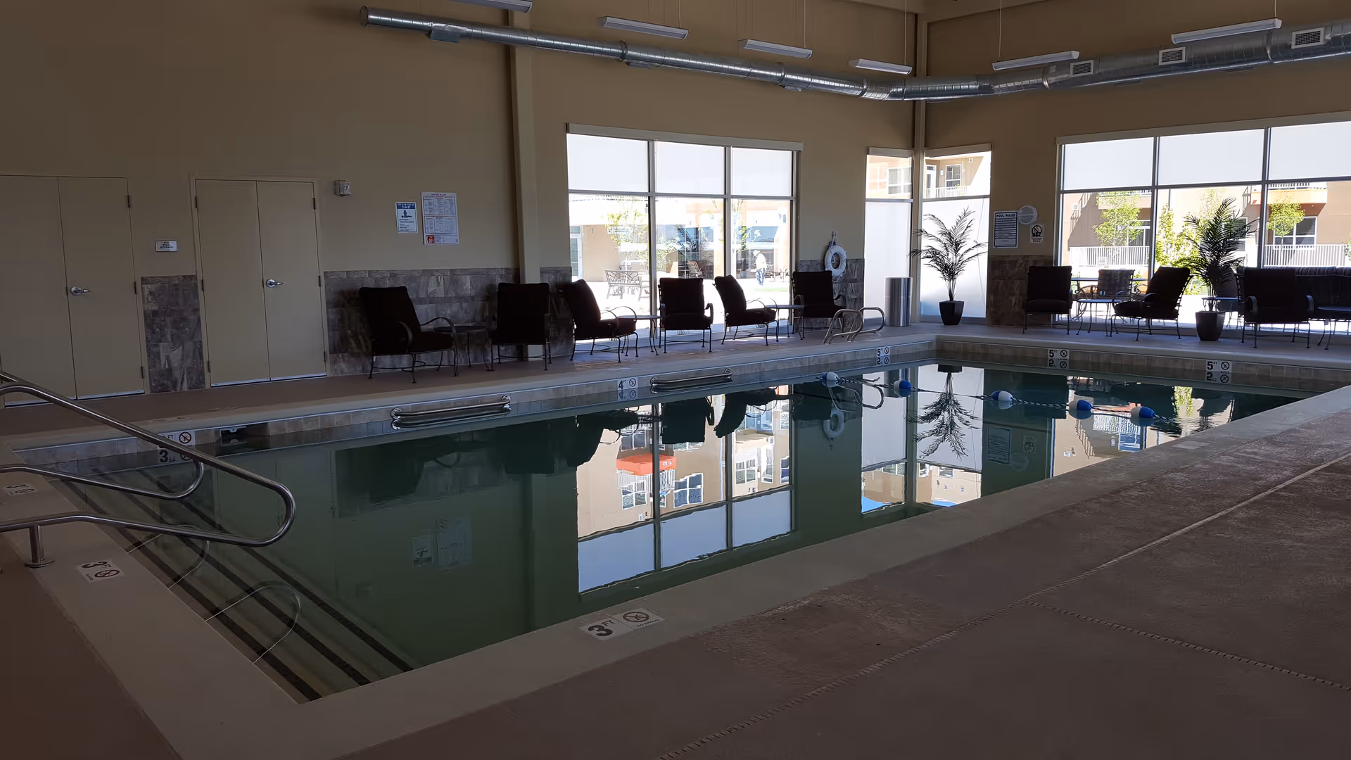 Indoor swimming pool area with large windows letting in natural light, several chairs and potted plants arranged along the walls, and a handrail leading into the pool.