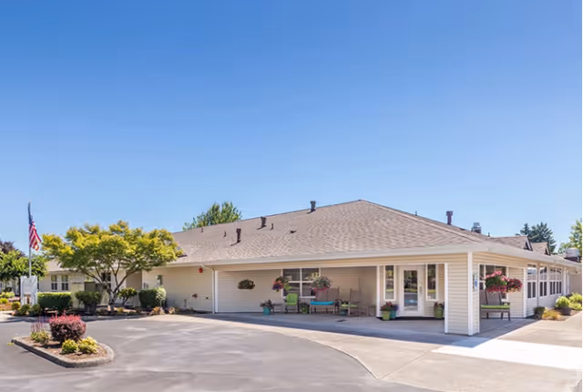 Exterior view of a single-story senior living facility building with beige siding, a gray roof, and several windows. There are potted plants and hanging flower baskets near the entrance, a small landscaped area with bushes and trees, and an American flag on a flagpole. The sky is clear and blue.