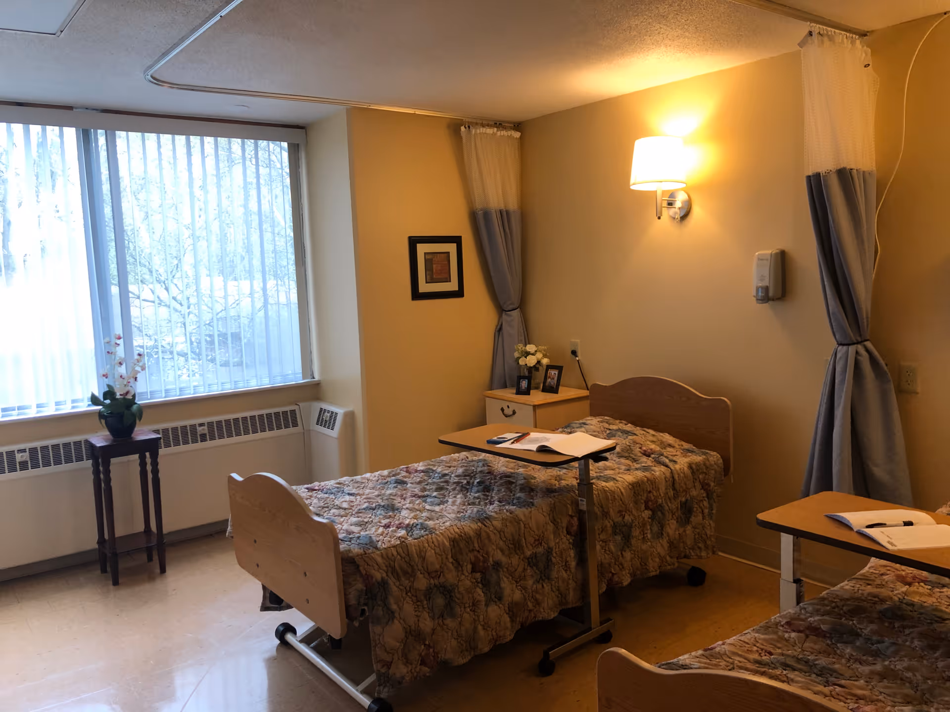A shared bedroom in a rehabilitation and health care center with two hospital-style beds covered with patterned quilts. Each bed has an overbed table with notebooks and pens. The room has a large window with vertical blinds, a small wooden table with a potted plant, a nightstand with framed photos and flowers, and wall-mounted light fixtures. Privacy curtains are partially drawn around each bed.
