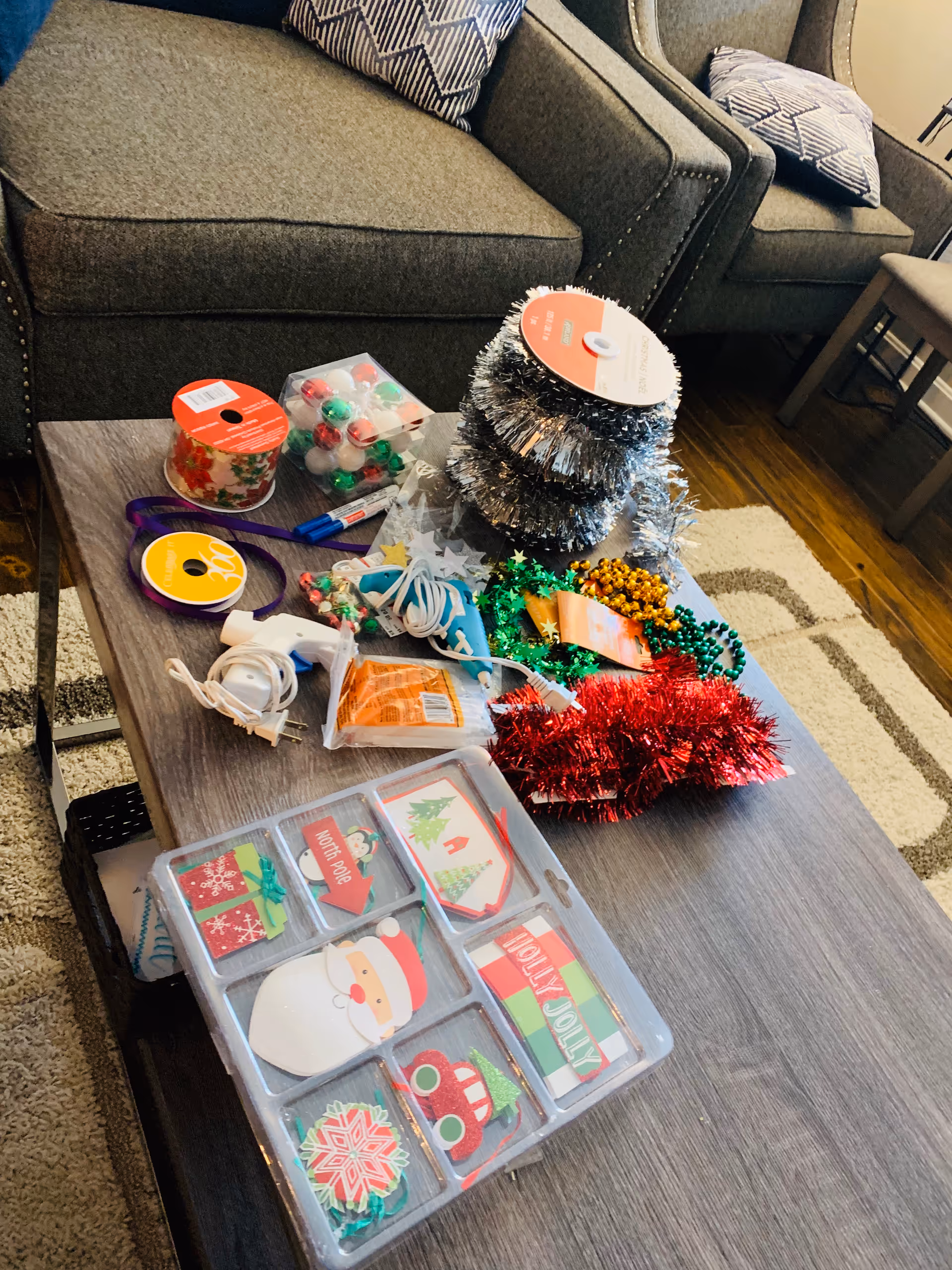 A living room coffee table covered with holiday decorations and craft supplies, with upholstered chairs and pillows in the background.