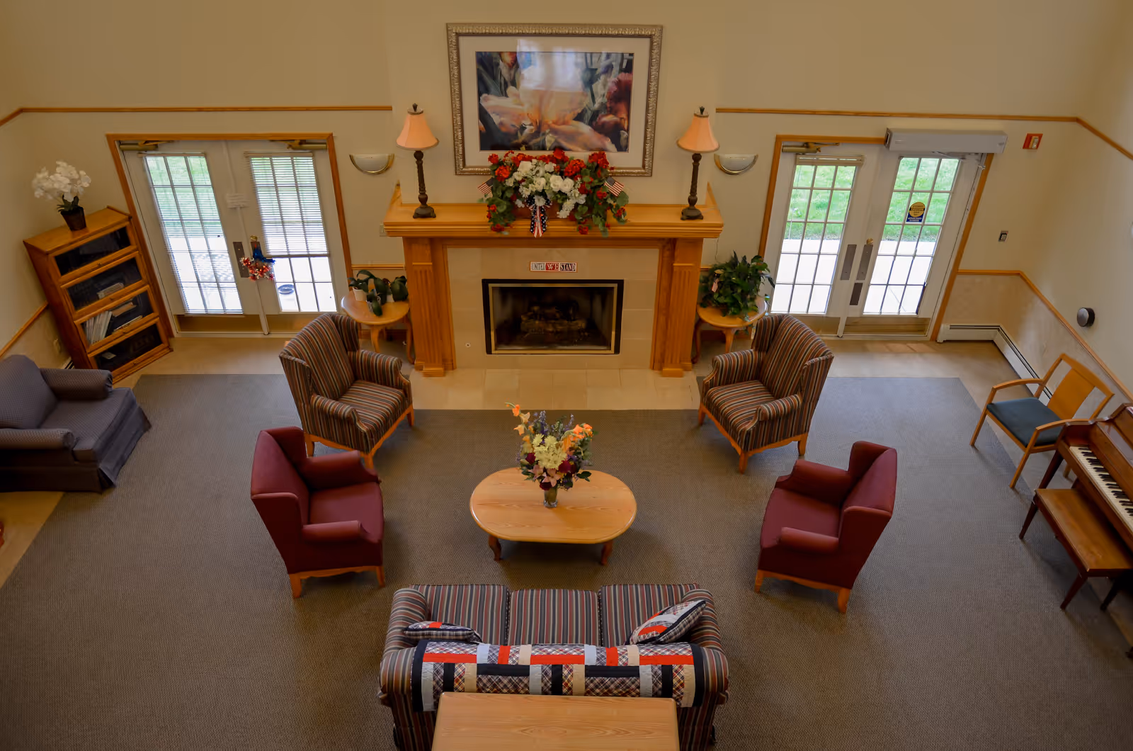 A cozy communal lounge with armchairs and sofas arranged around a central coffee table and fireplace, flanked by double doors, a piano, and bookshelves.