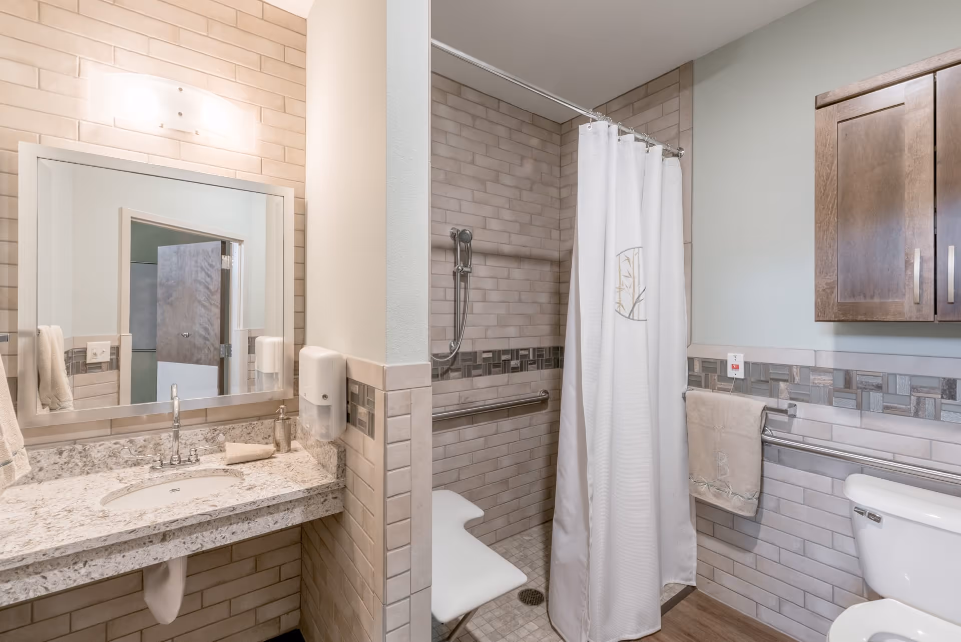 A clean and modern bathroom featuring a granite countertop with a sink, a large mirror above the sink, a wall-mounted soap dispenser, and a towel hanging on the side. The shower area has beige tiled walls, a white shower curtain with a decorative emblem, a handheld showerhead, and a white shower chair. There is a wooden cabinet mounted on the wall above the toilet, and the walls are decorated with a tile border.