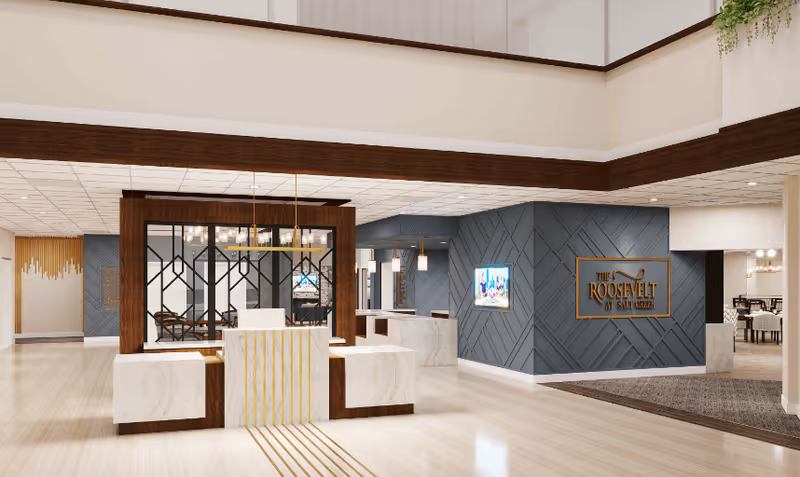 Spacious and modern lobby area of The Roosevelt at Salt Creek featuring a central reception desk with marble and wood accents, a decorative partition with geometric patterns, blue textured walls, a wall-mounted TV, and a sign displaying the facility's name. The area is well-lit with ceiling lights and has light-colored flooring.
