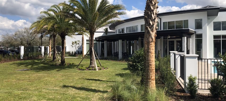 Outdoor view of a senior living facility with a well-maintained lawn, several palm trees, and a modern building with large windows and a covered patio area under a partly cloudy sky.