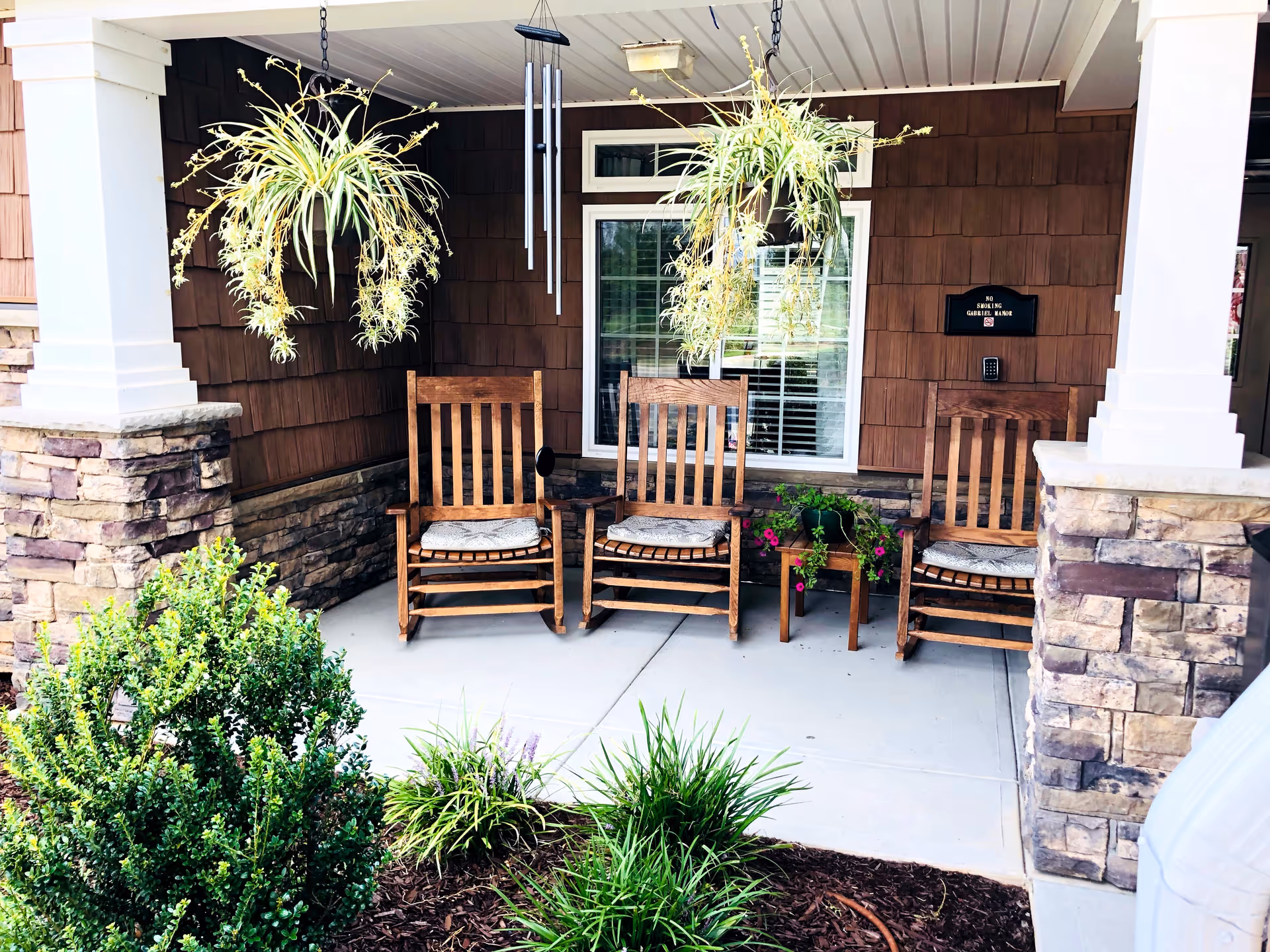 A covered porch area with three wooden rocking chairs with cushions, two hanging plants, a small wooden table with a potted plant, and a window with white blinds. The porch has stone pillars and brown shingle siding. There is a 'No Smoking Gabriel Manor' sign on the wall.
