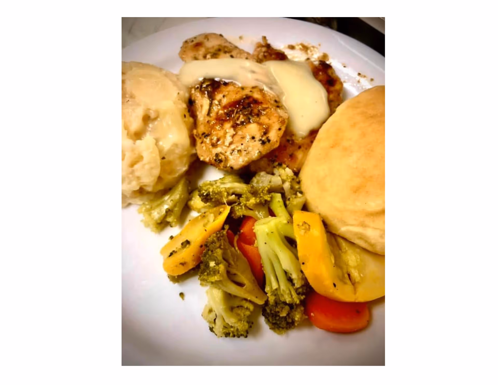 A plate with grilled chicken topped with sauce, mashed potatoes, a biscuit, and mixed vegetables (broccoli, carrots, yellow squash).