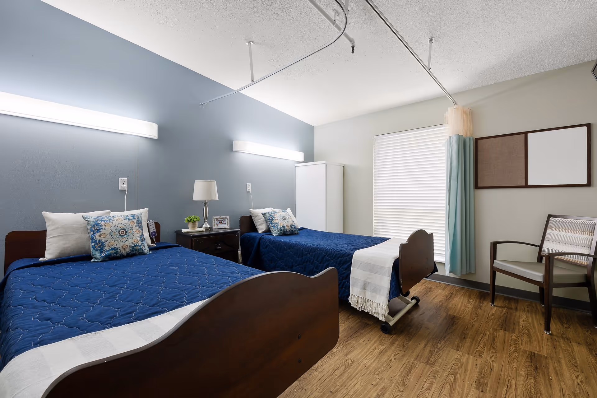 A nursing facility bedroom with two single beds covered in blue quilts and white pillows. Between the beds is a small nightstand with a lamp, a small plant, and a framed photo. The room has light blue and white walls, a window with blinds and a curtain, a wooden chair, and a white cabinet. The floor is wood-patterned.
