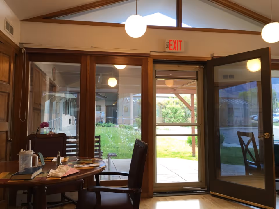 Interior view of a room with wooden furniture including a round table and chairs. The room has large glass doors and windows looking out to a green outdoor area. A door is open leading outside. Ceiling lights are visible and there is an exit sign above the door.