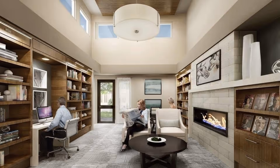 A bright and modern senior living common area with high ceilings and large windows. The room features built-in wooden bookshelves filled with books and decorative items on both sides. On the left, a man is seated at a desk using a computer. In the center, a woman is sitting in a comfortable chair reading a newspaper, and another woman is seated at a table in the background. On the right side, there is a contemporary fireplace with a framed horse artwork above it and magazines displayed on shelves below. A round coffee table with a decorative plate is in the middle of the room.