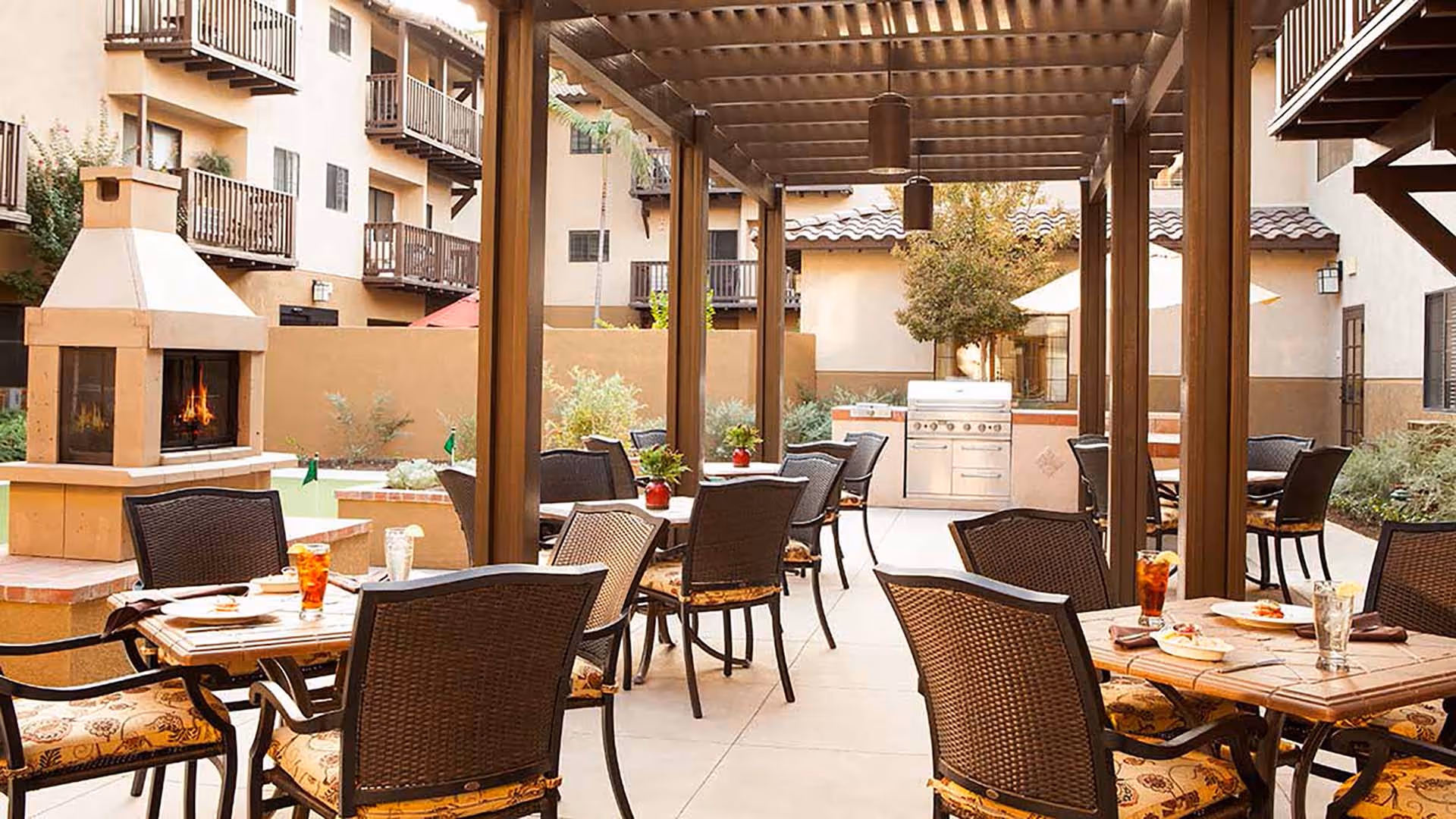 Outdoor patio area at Atria Del Rey with multiple tables and chairs under a wooden pergola, a built-in fireplace with a fire burning, and a stainless steel grill in the background. The space is surrounded by a beige building with balconies and some greenery.