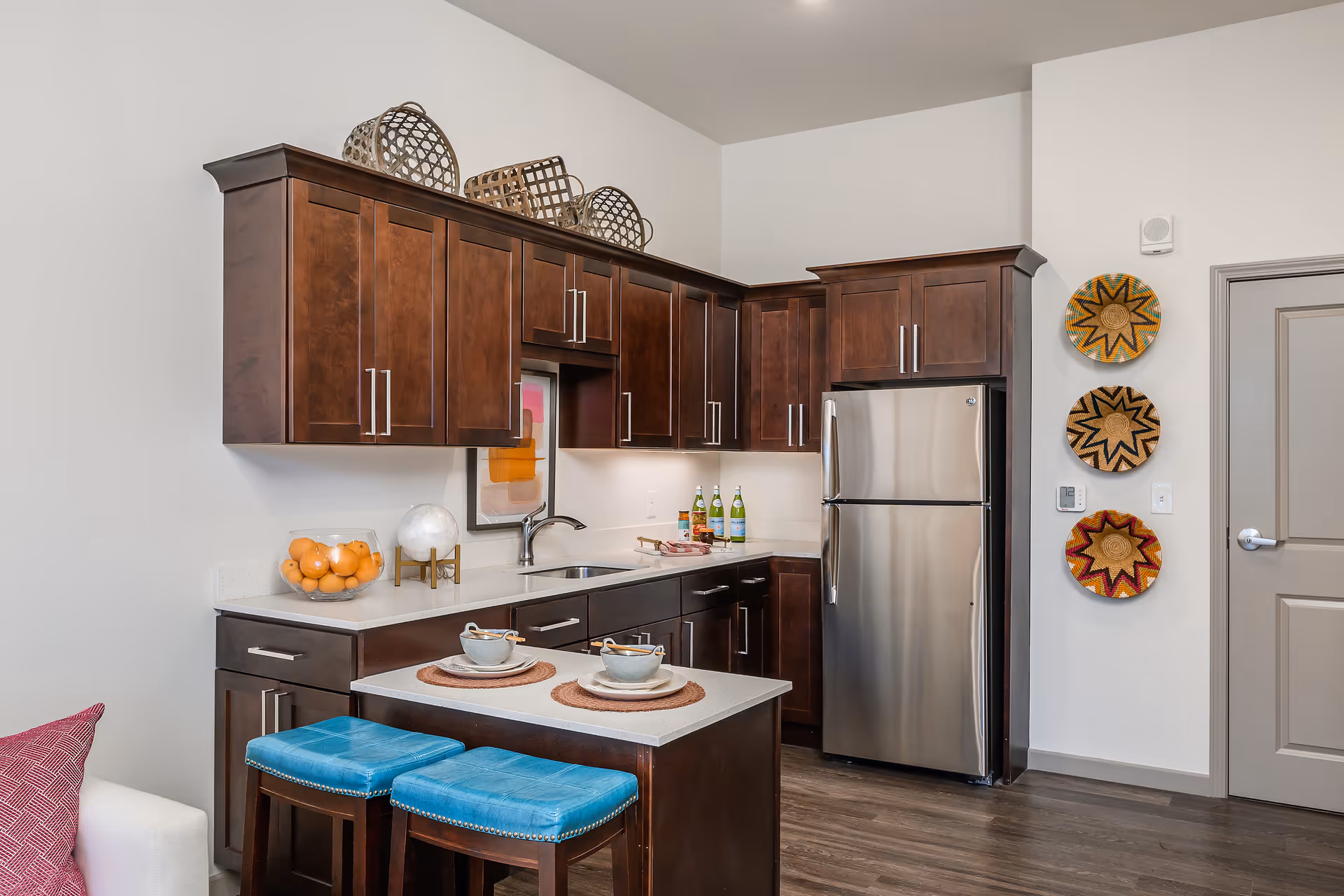 Modern kitchen area with dark wooden cabinets, a stainless steel refrigerator, a small island with two blue cushioned stools, and decorative baskets on the wall. The countertop has a bowl of oranges, a globe, and two place settings with cups and plates.