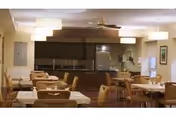 A dining room with multiple tables and chairs arranged neatly. The room features warm lighting with ceiling lamps and a ceiling fan. In the background, there is a kitchen area with a refrigerator and cabinets.