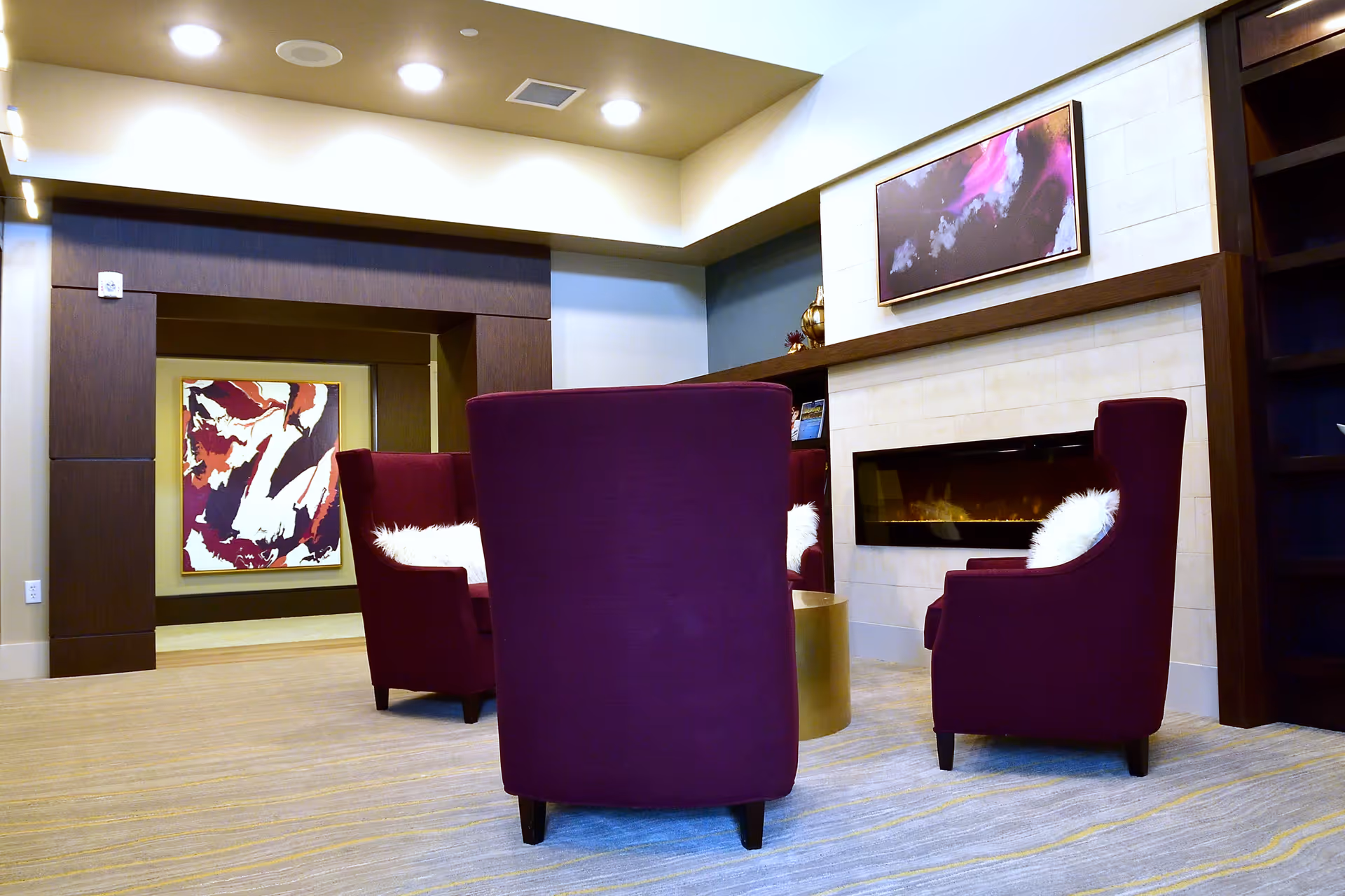 A cozy senior living common area with three purple armchairs arranged around a small round table. The chairs have white fluffy pillows. There is a modern electric fireplace built into a light-colored tiled wall, above which hangs an abstract painting with purple and pink hues. The room has recessed lighting and a carpeted floor with subtle patterns. In the background, there is a dark wood-framed opening leading to another area with a colorful abstract painting on the wall.