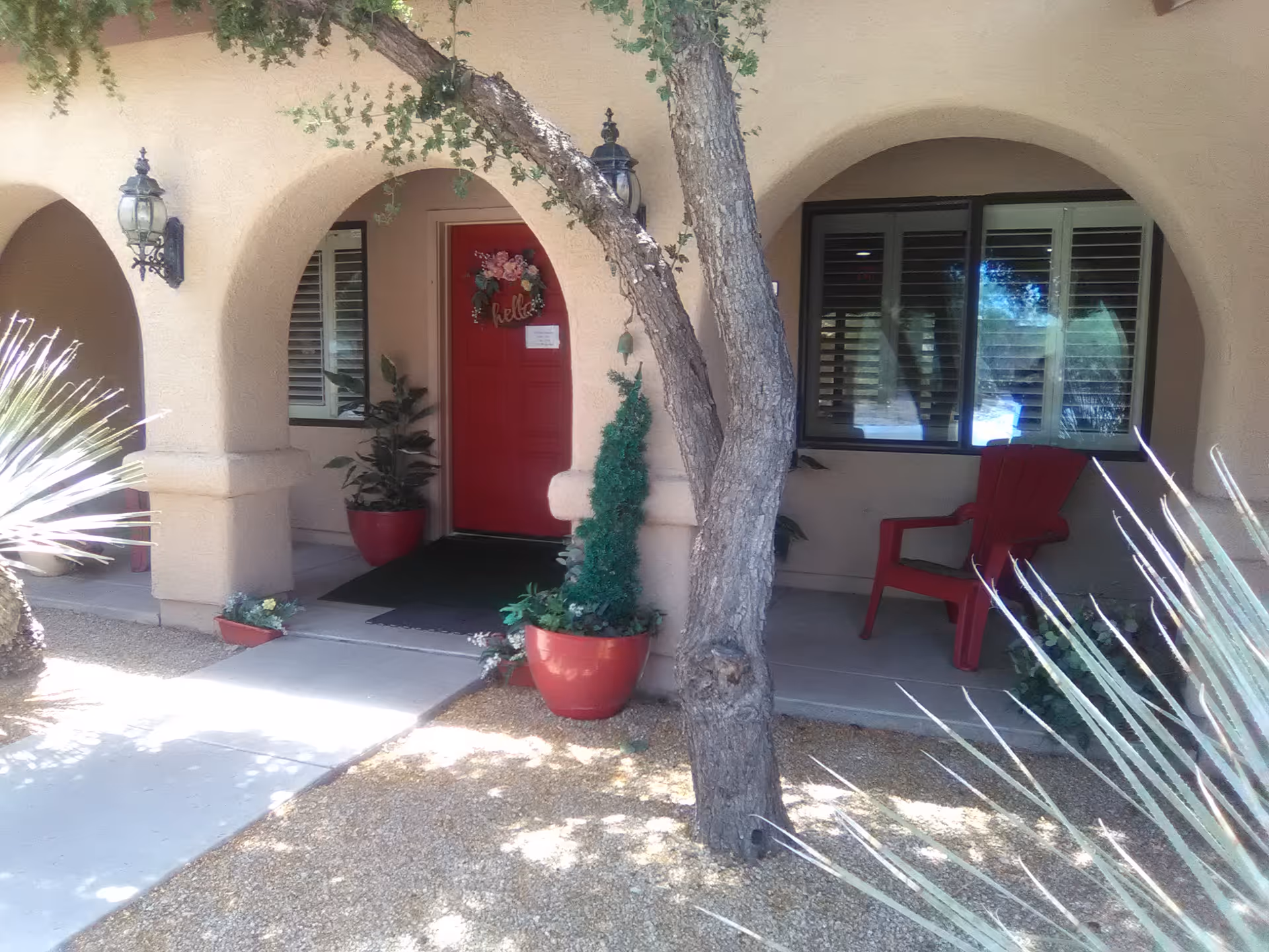Front porch of a stucco building with a red door, potted plants, an arched entry, and a tree in the foreground.