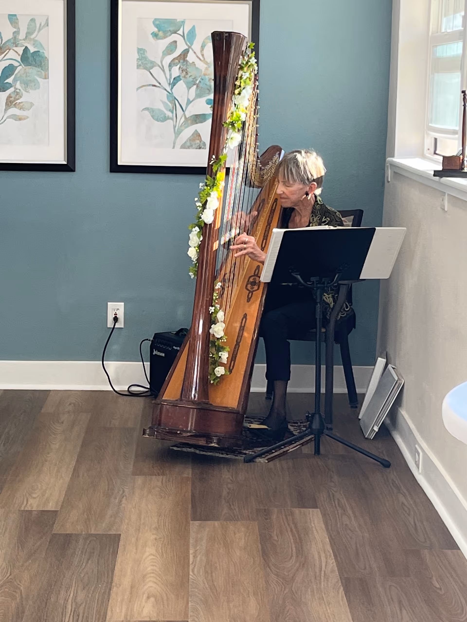 An elderly woman seated indoors playing a large wooden harp decorated with white flowers. She is positioned near a music stand with sheet music, against a teal wall with two framed botanical prints. The room has wood flooring and a window letting in natural light.