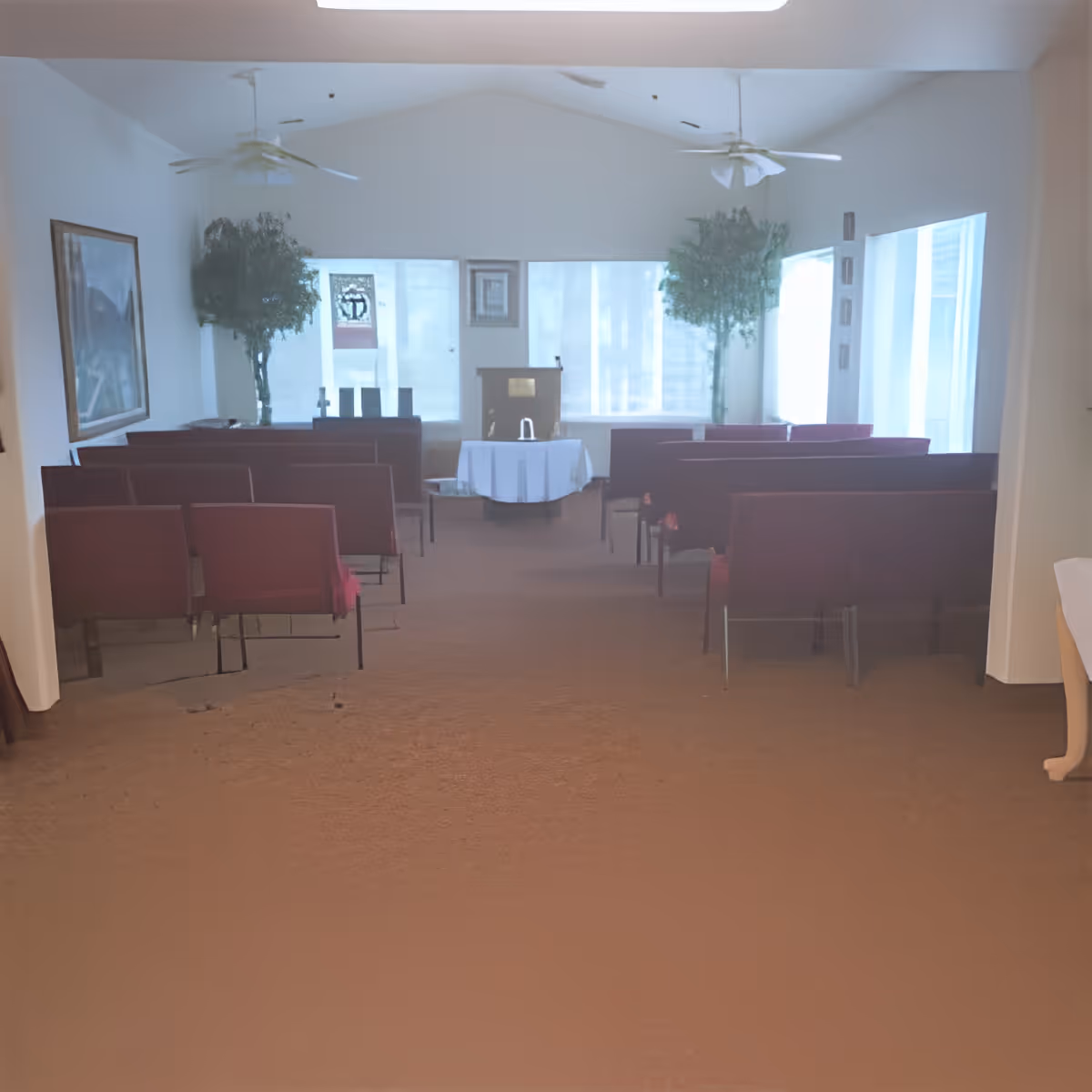 Interior view of a small chapel or meeting room with rows of red chairs facing a podium and a table covered with a white cloth. The room has large windows with sheer curtains, two ceiling fans, and potted plants on either side of the podium. There is a framed picture on the left wall.