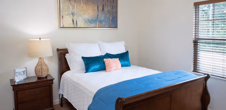A neatly made bed with white bedding, two teal pillows, and one pink pillow in the center. The bed has a wooden frame and a blue throw blanket at the foot. To the left of the bed is a wooden nightstand with a woven lamp and a framed sign that says 'rise up & PRAY'. A large abstract painting hangs on the wall above the bed. To the right, there is a window with wooden blinds partially open, letting in natural light.
