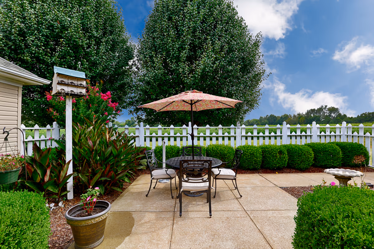 Outdoor patio area with a round table and four chairs under a patterned umbrella, surrounded by green bushes, trees, and a white picket fence under a partly cloudy blue sky.